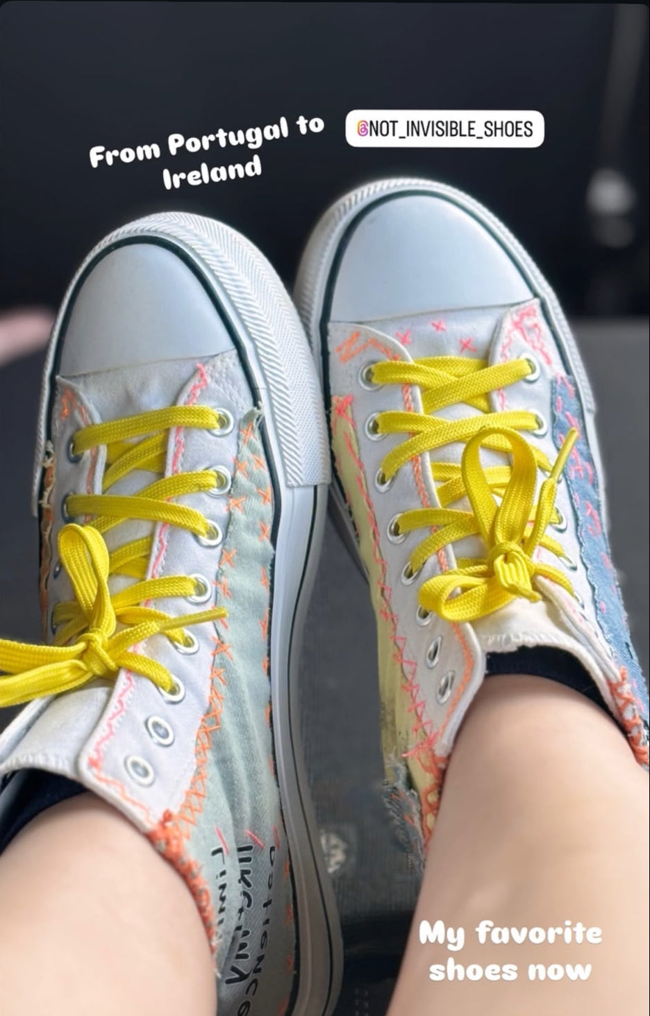 A pair of high-top canvas sneakers worn on feet, shown in a social media-style post. The shoes have a light gray base with pastel multicolor paint splatter, bright yellow laces, and black socks.